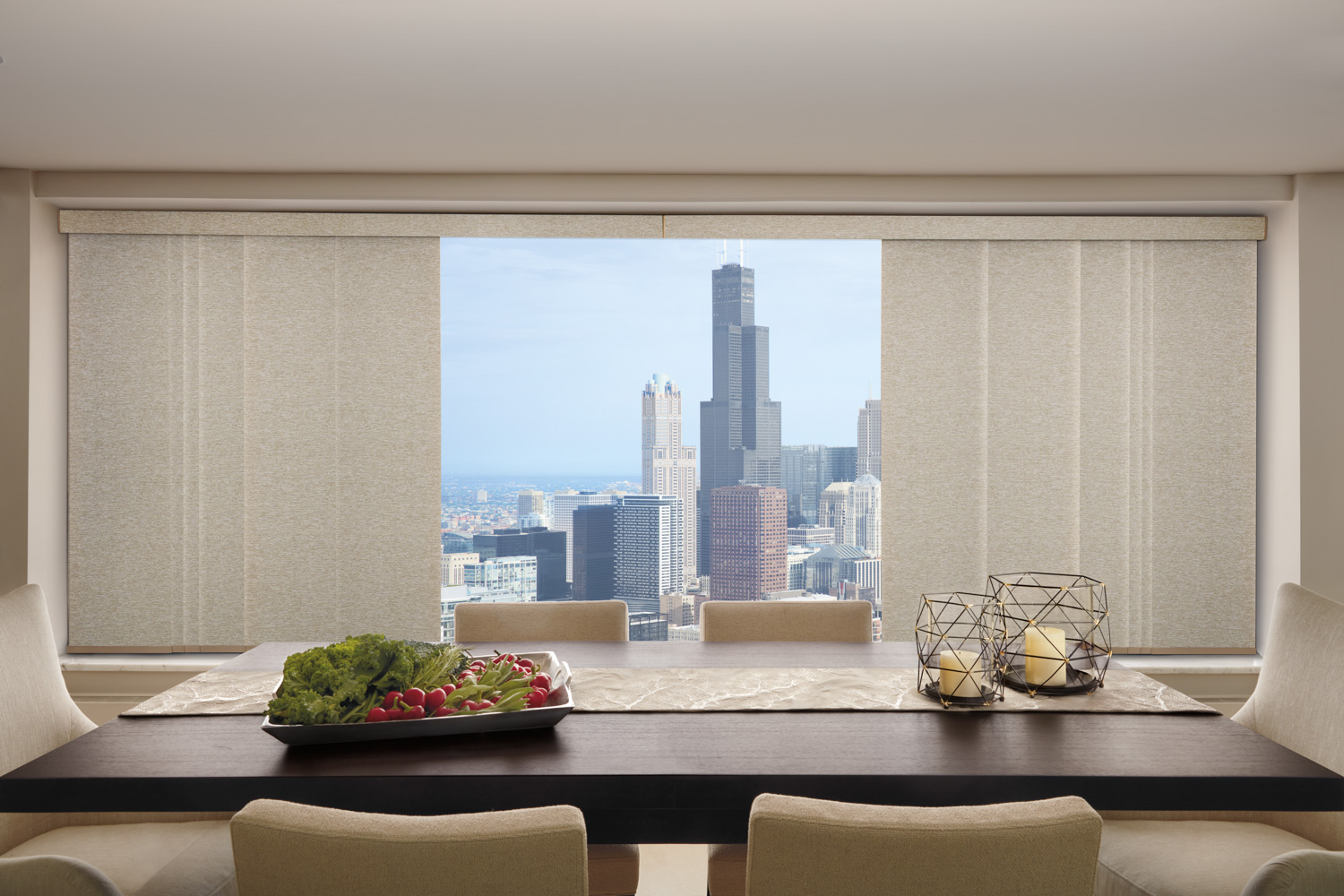 A dining room with a table and chairs in front of a window, showcasing a city view. Hunter Douglas Skyline® Panel-Track Blinds.
