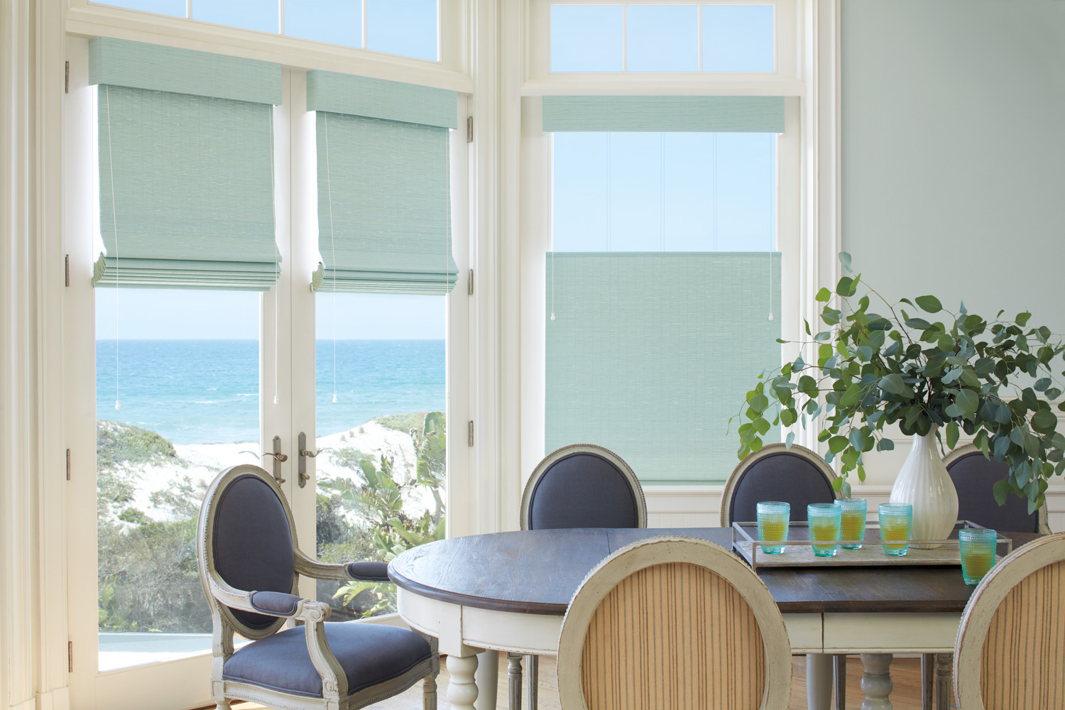 A dining room with a table and chairs in front of an ocean-view window, adorned with Hunter Douglas Provenance® Woven Wood Shades.