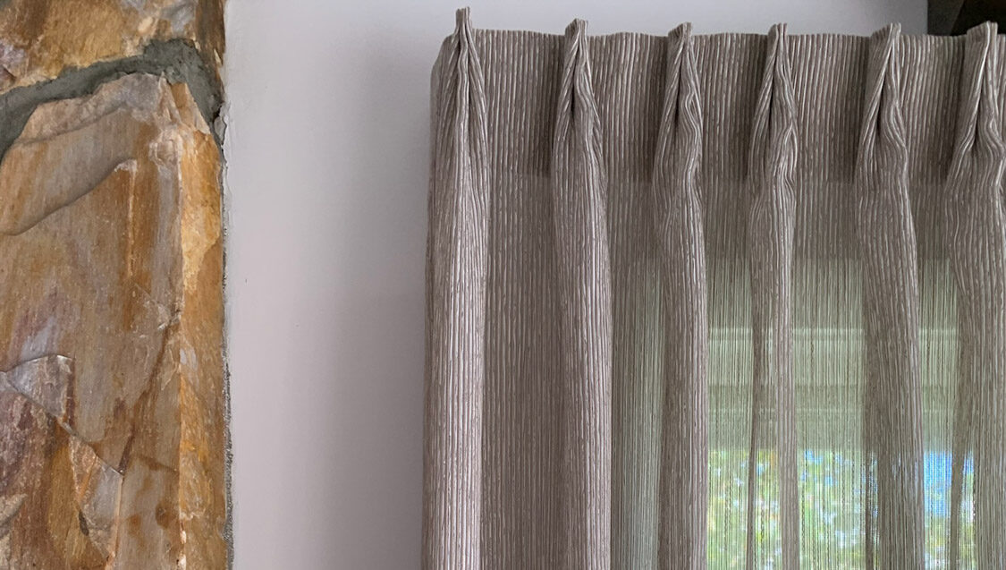 Sheer curtains with Inverted Pleat cascading in front of a stone wall.